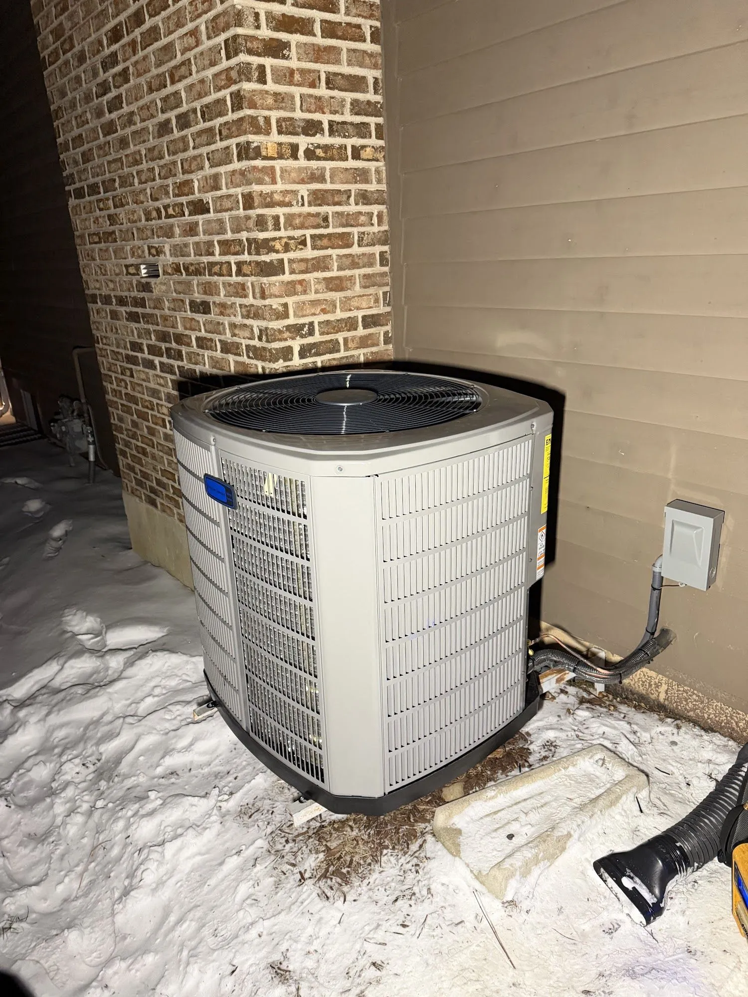 An outdoor condensing unit standing in snow beside a brick wall, photographed at night with a flash.