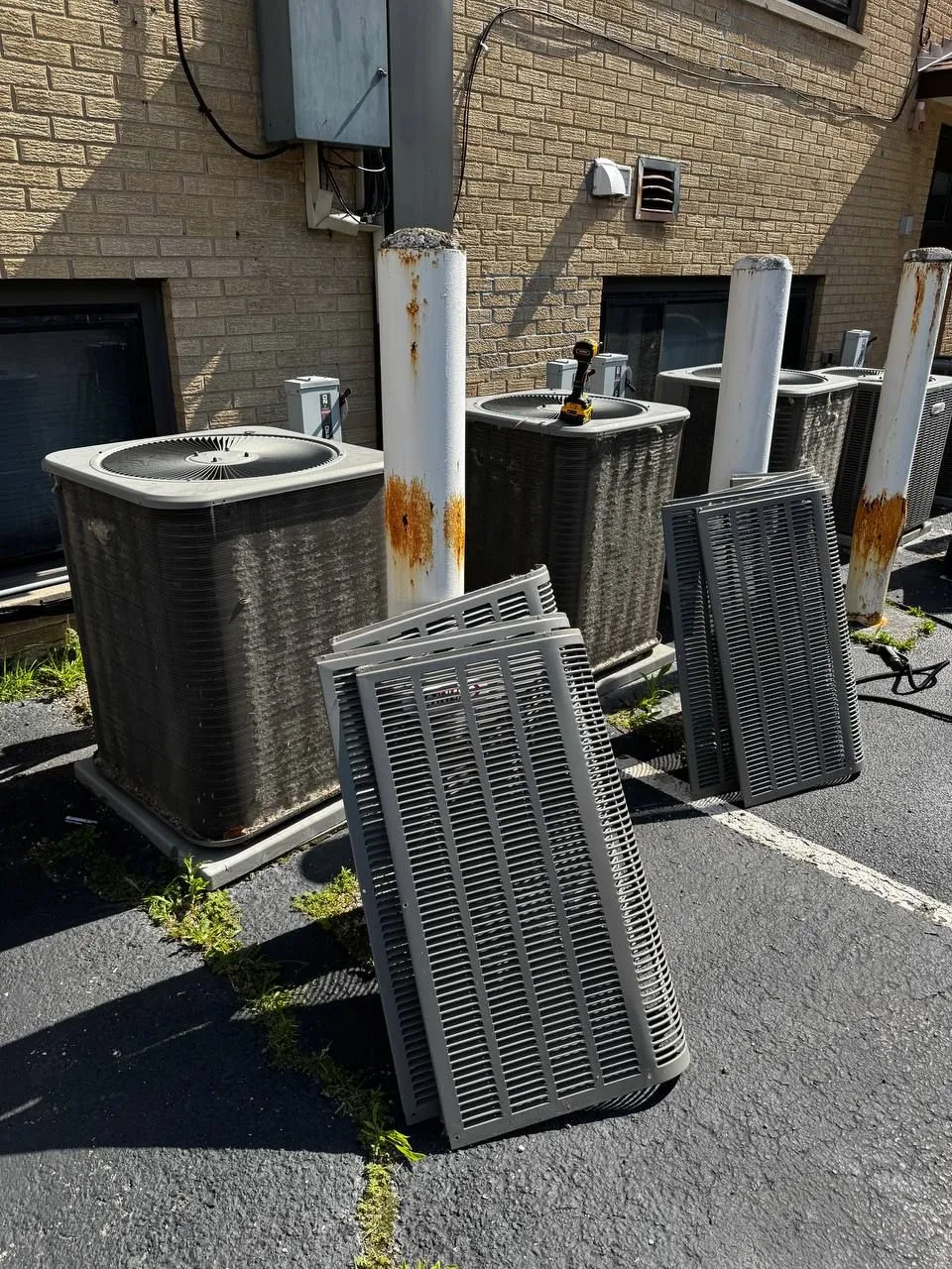 A row of outdoor condensing units beside a tan brick building, their louvered side panels taken off and stacked on the asphalt.