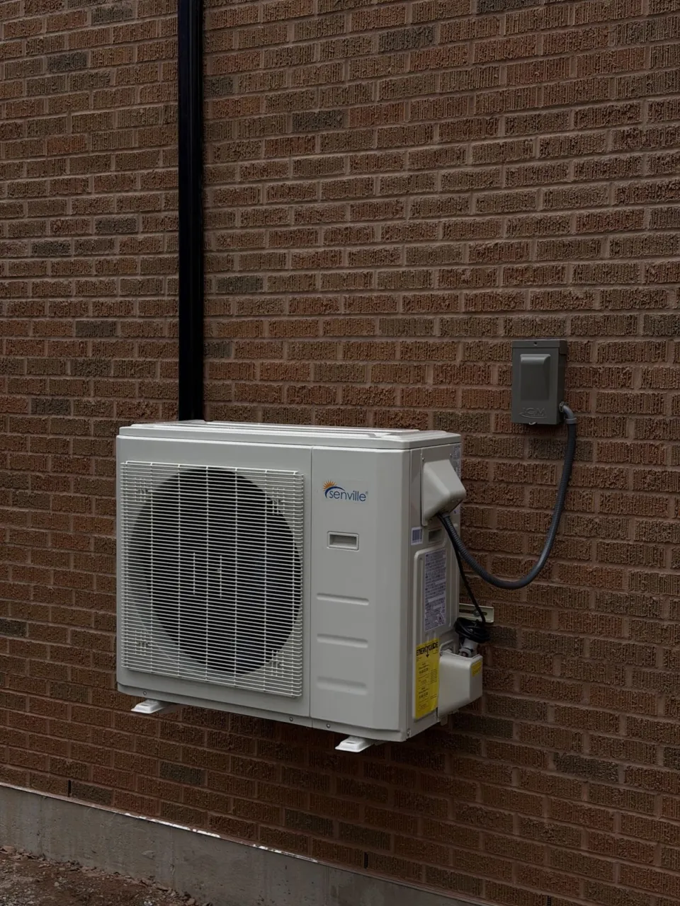 A ductless outdoor unit mounted on wall brackets above the ground against dark brick, with its disconnect beside it.