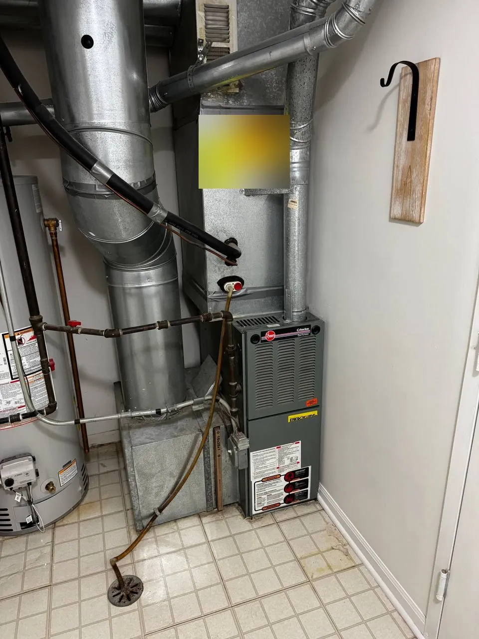 An old Rheem gas furnace and its venting in a small tiled utility closet next to a storage tank, before removal.