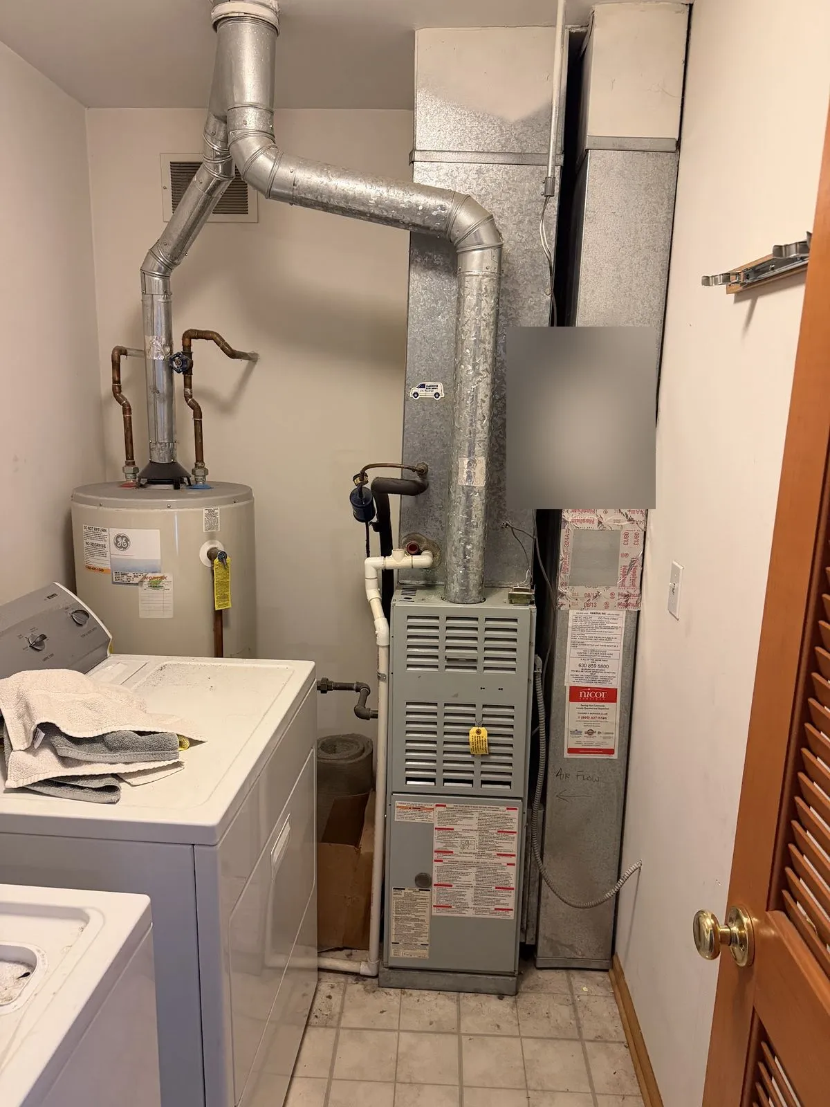 An old gas furnace in a laundry closet beside a washer and a storage tank, before replacement.