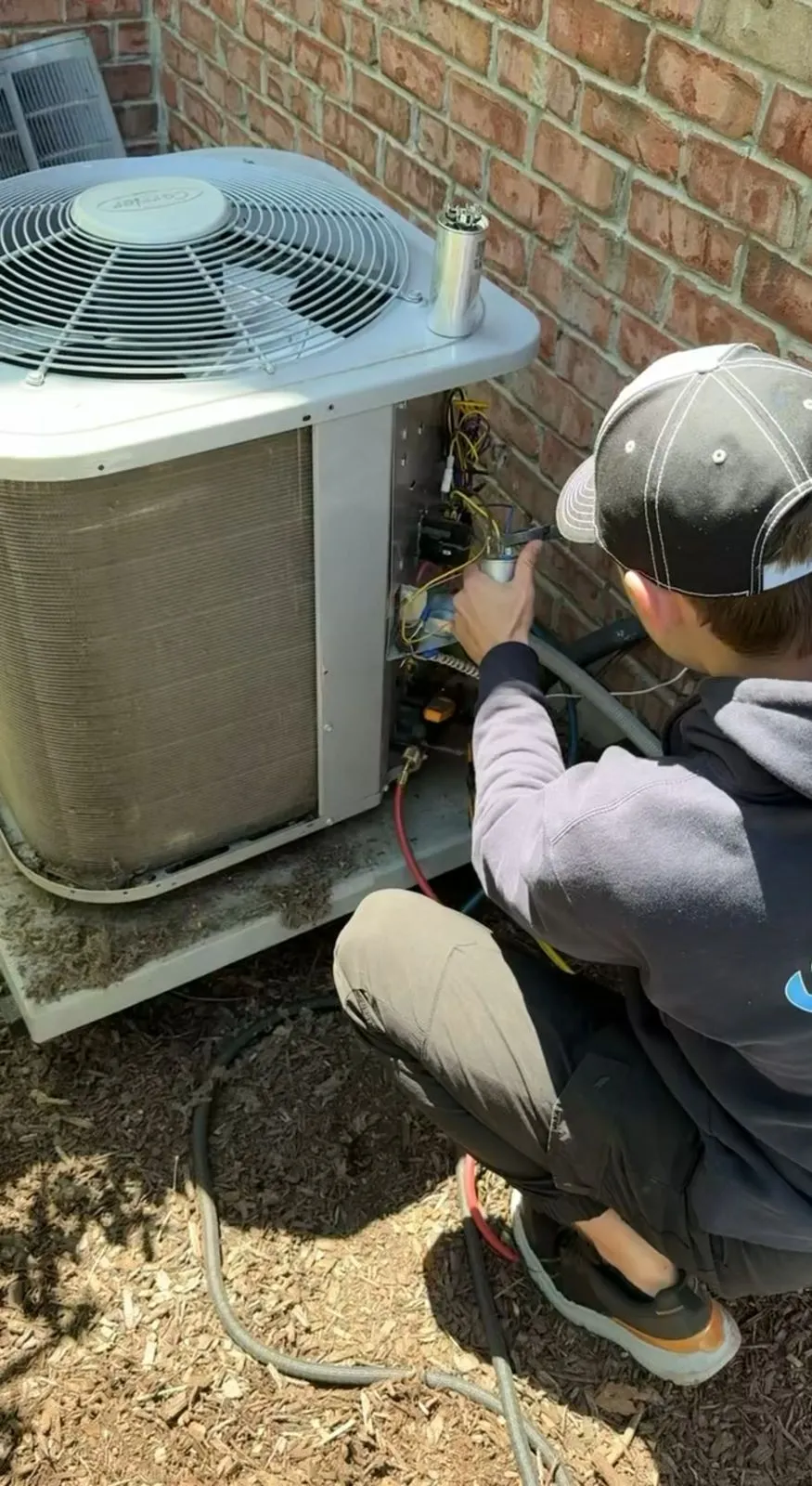 A technician crouched at the open service panel of an outdoor air conditioner, testing the wiring, with a capacitor standing on top of the unit.
