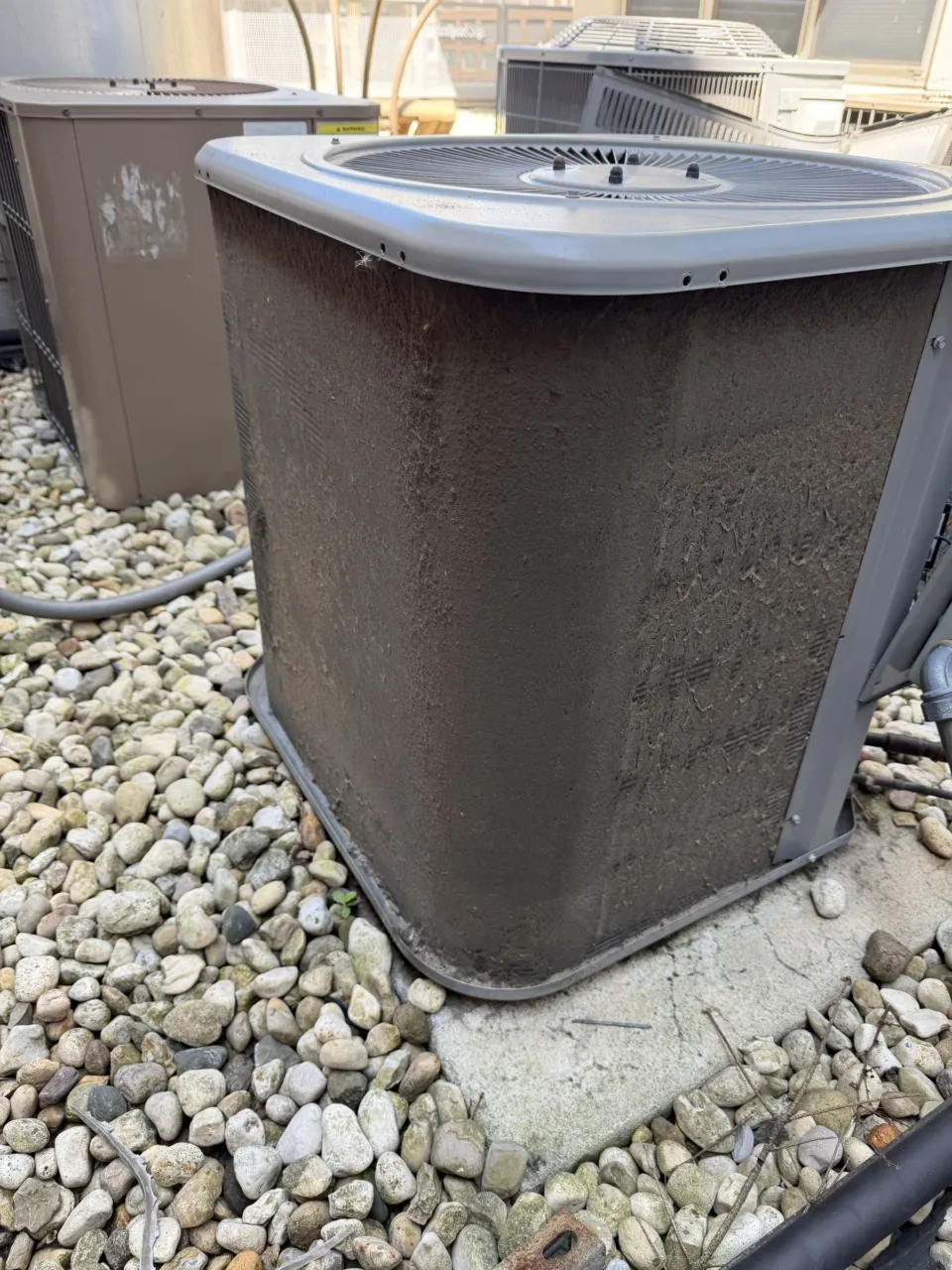 Outdoor air conditioner with its condenser coil packed solid with dirt and cottonwood, standing on a gravel bed.