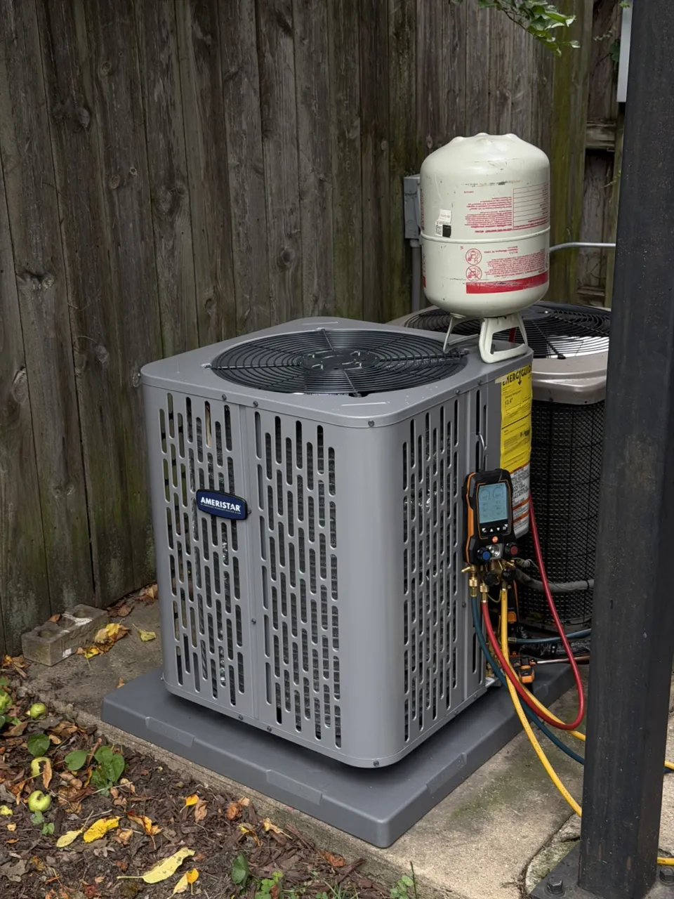 A new outdoor condensing unit on a fresh pad in the same spot by the fence, gauges still connected during start-up.