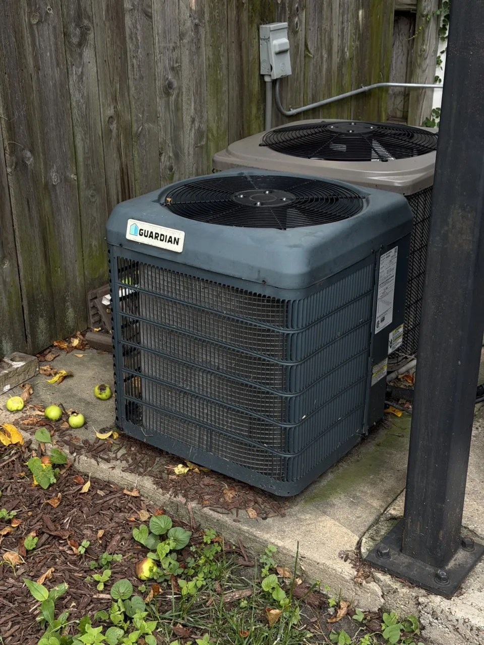 An old outdoor condensing unit standing beside a wooden fence, rust on the casing.