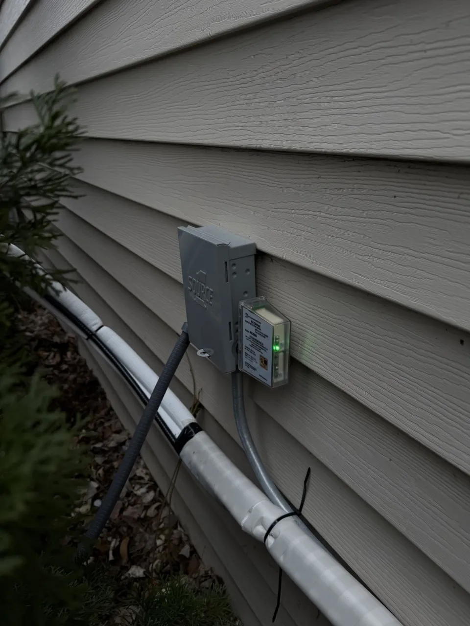 An outdoor electrical box and an Intermatic surge protector with its green indicator lit, mounted on beige siding above the white-wrapped line set.