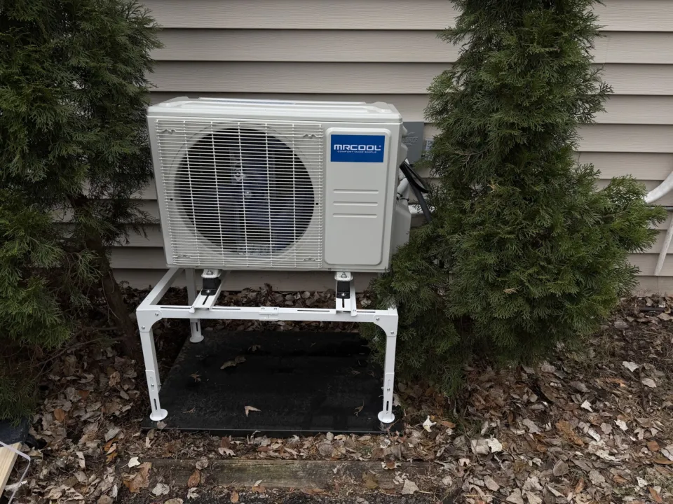 A MrCool outdoor unit on a white floor stand and pad between two evergreen shrubs, against beige siding.