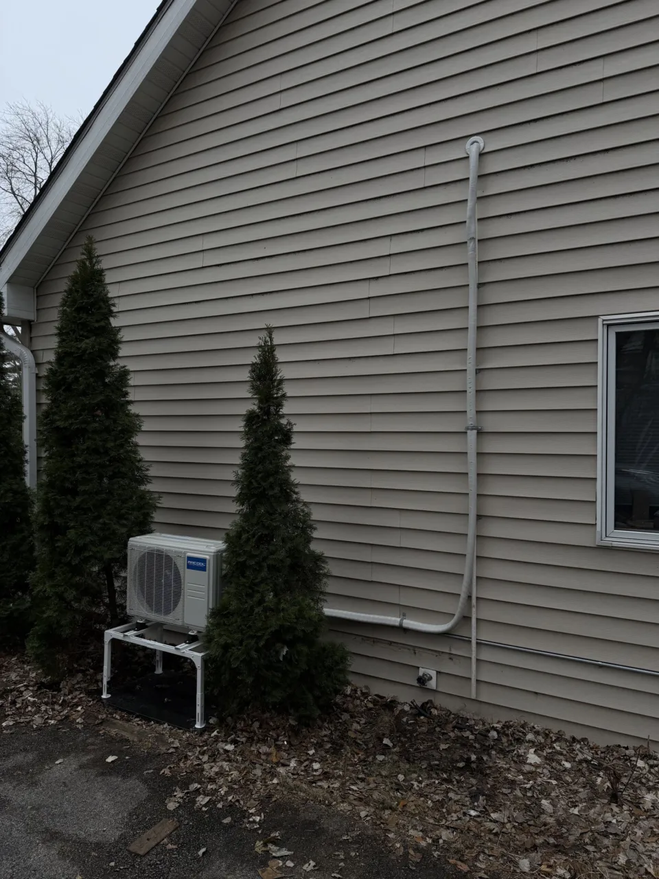 The side of a house with beige siding, the new outdoor unit on its stand between evergreens and the line set cover running up and along the wall.