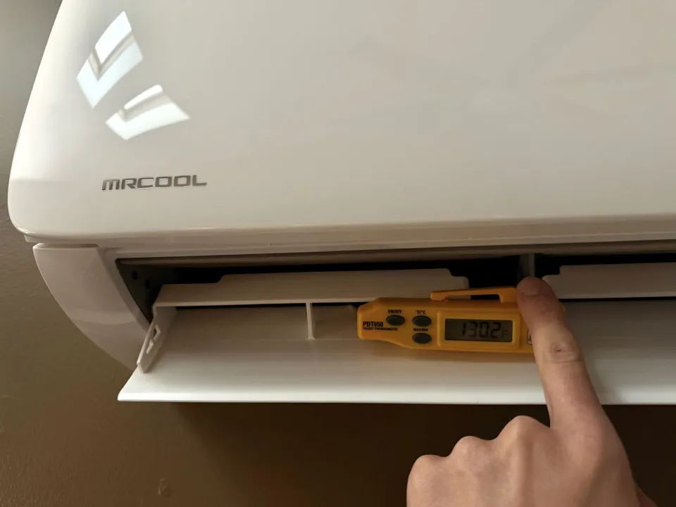 A pocket thermometer held in the air outlet of the MrCool indoor unit, reading 130.2 F.