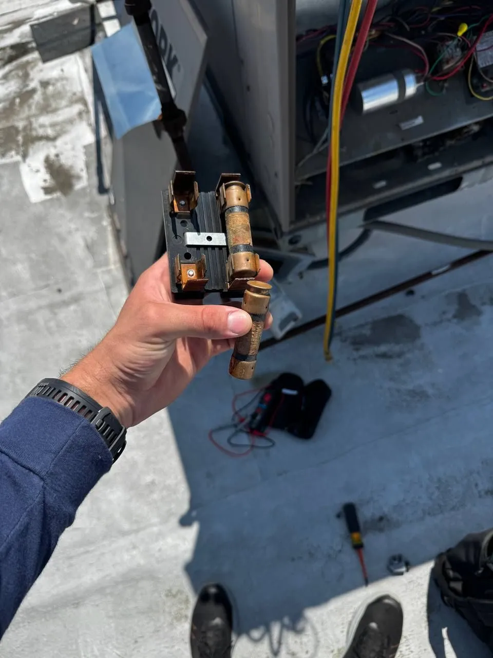 A hand holding a fuse block and a scorched cartridge fuse taken out of a rooftop unit, the open unit and test leads on the roof behind.