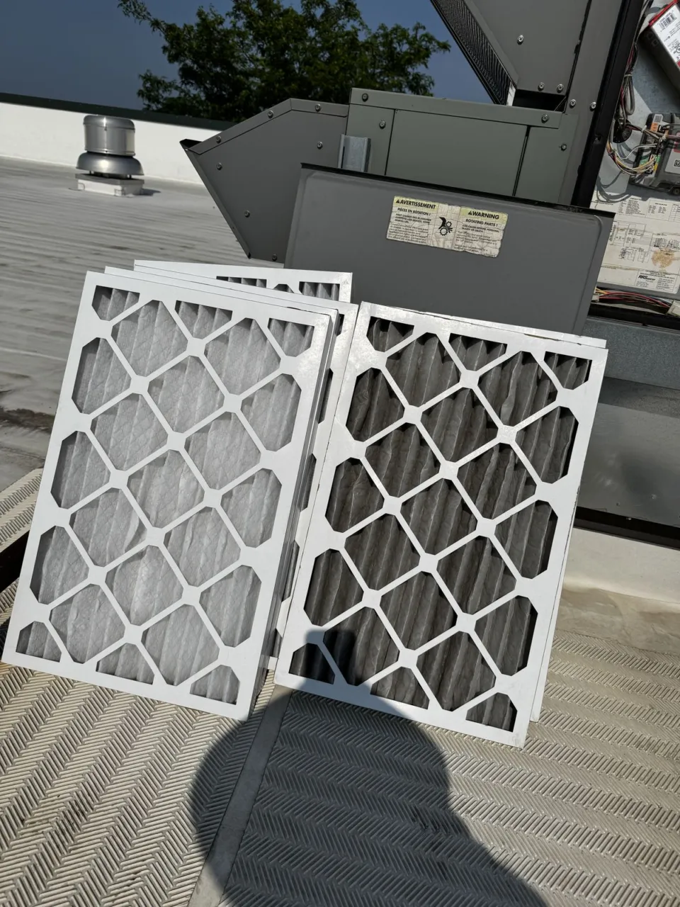 Two stacks of pleated air filters leaning against a rooftop unit on a flat commercial roof, the right-hand stack gray with dust.