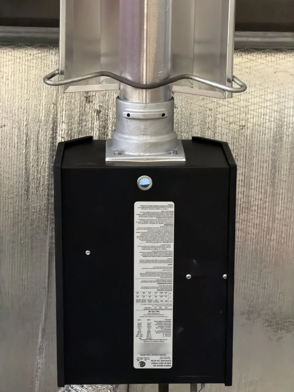 Close-up of the black burner box at the end of a radiant tube heater, its flue collar bolted to the tube and the stainless reflector above it.