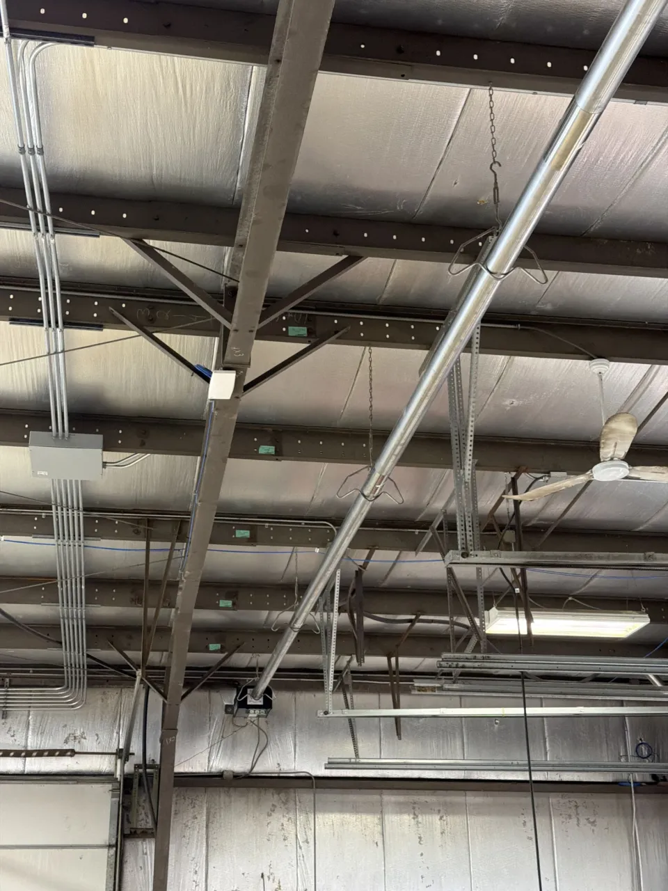 A galvanized gas-fired infrared tube heater hung on chains from the roof steel of a warehouse bay, running diagonally beneath the insulated deck.