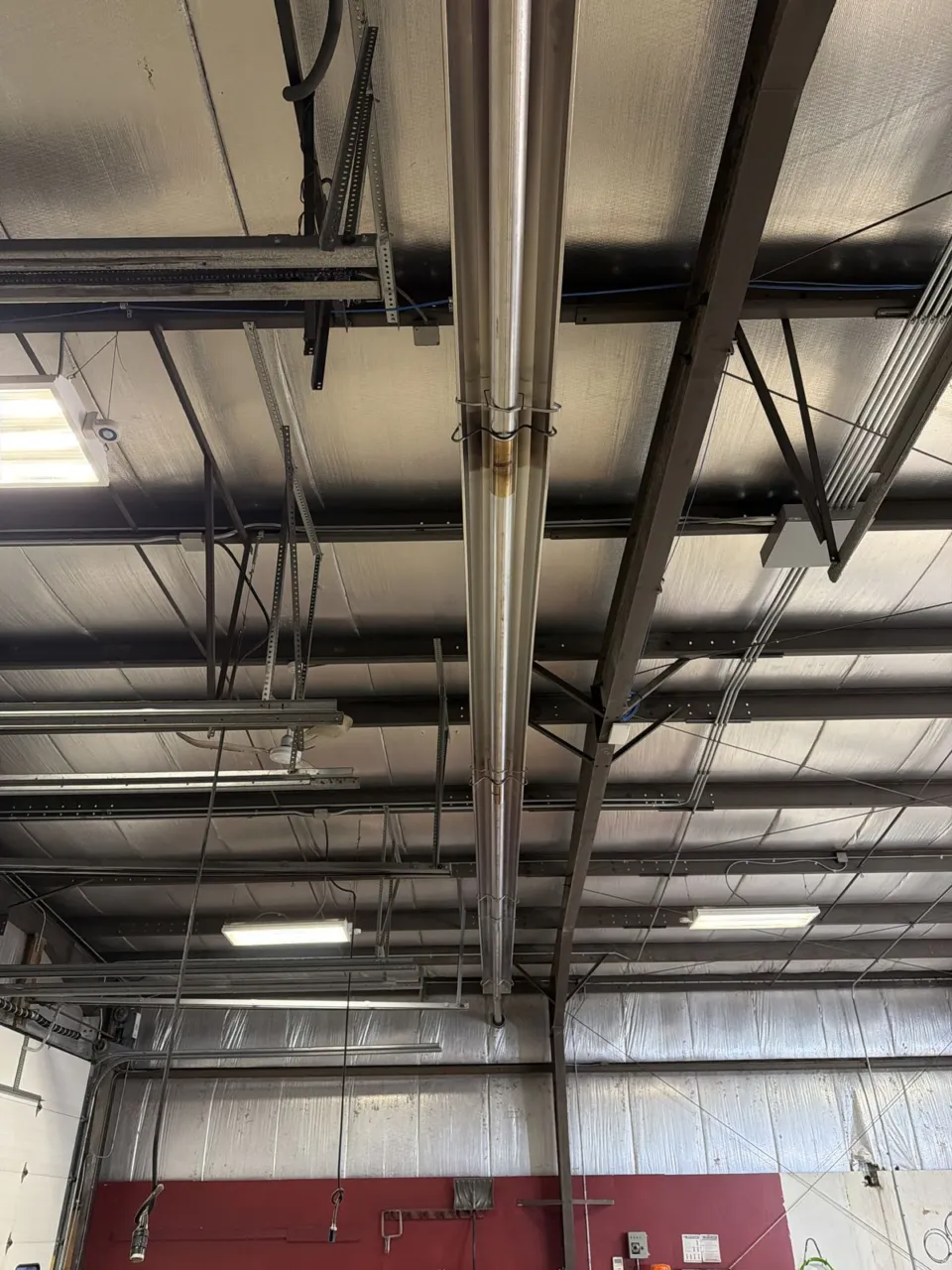 A second radiant tube heater run above a warehouse bay, hung between the roof joists and the strip lights, with the painted wall of the bay below.