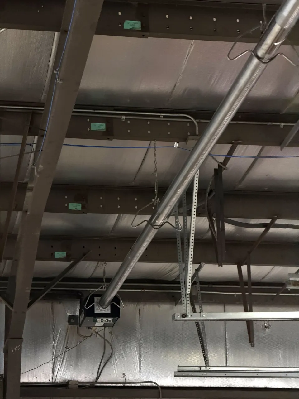 The same tube heater seen along its length between the roof joists, dropping to the black burner box bolted to the wall at the end of the run.