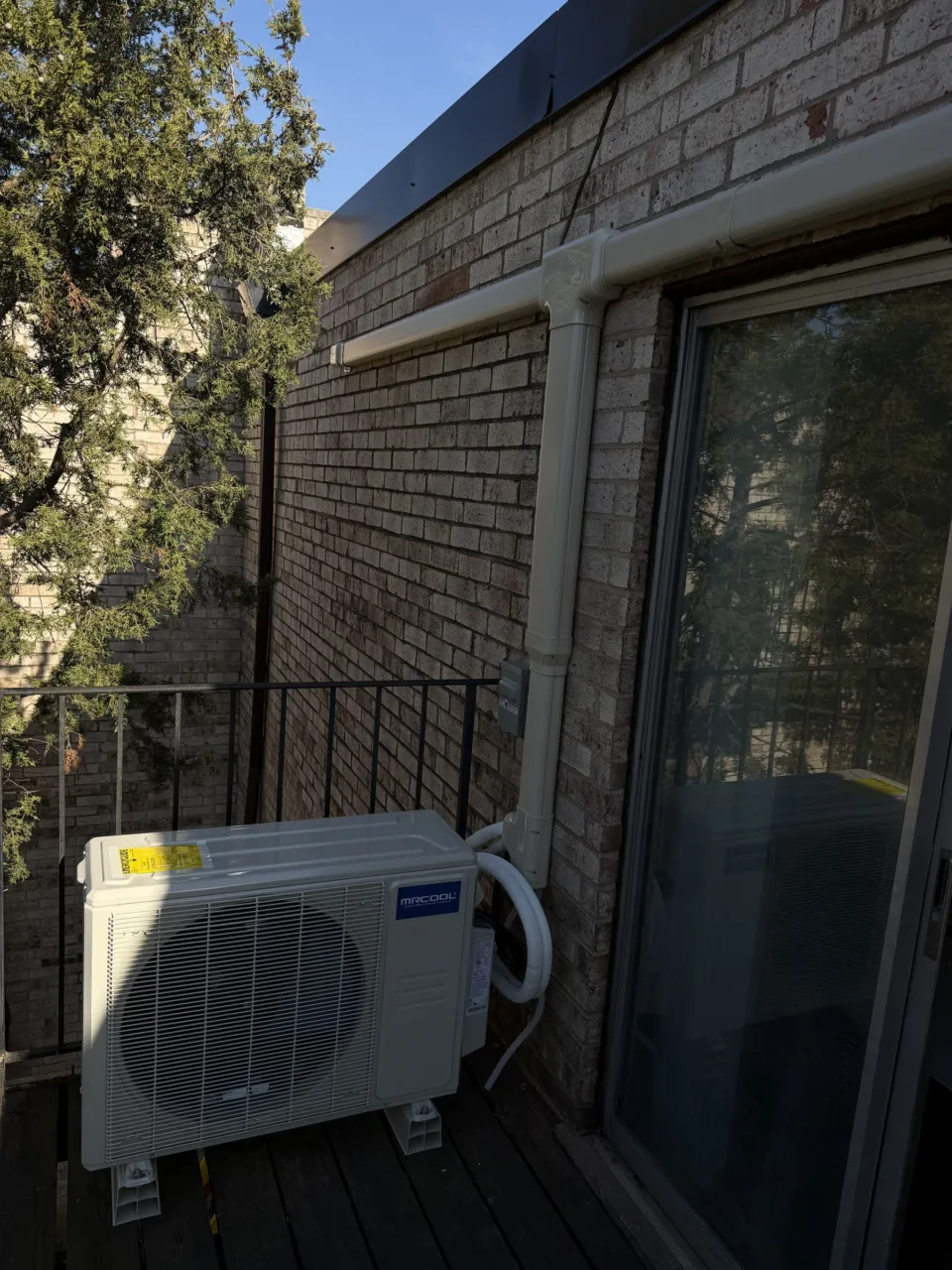 A MrCool multi-zone outdoor unit standing on feet on a wooden balcony deck beside a light brick wall, its white line set cover running up the wall and along the soffit.