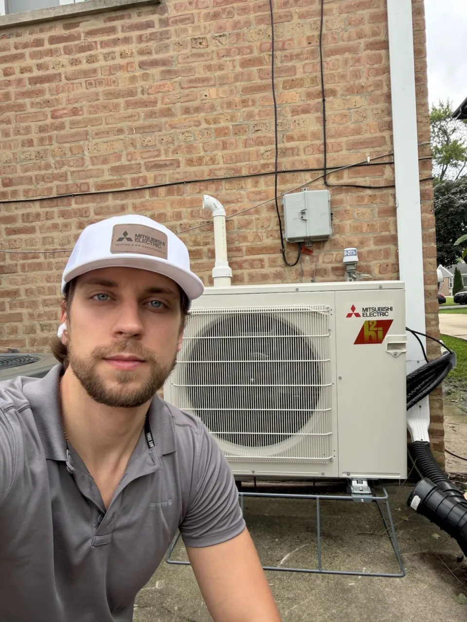 Alex, the owner of General Climate, in a white Mitsubishi Electric cap next to a Mitsubishi Electric cold-climate heat pump on a brick wall.
