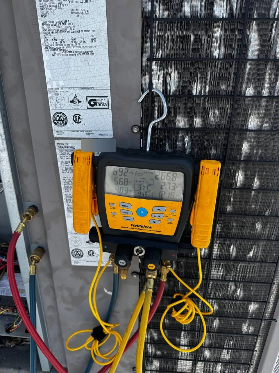 A Fieldpiece digital refrigerant manifold hung on a packaged outdoor unit next to its rating plate, showing pressure and temperature readings.